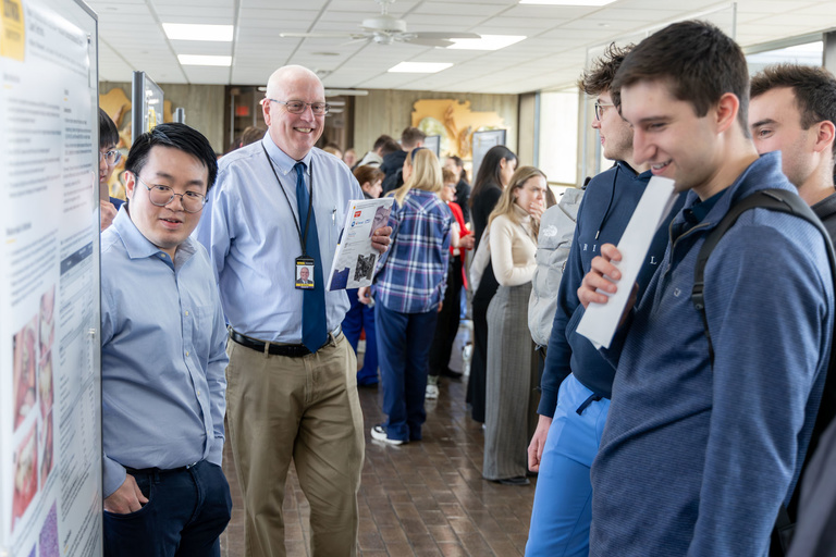 A student talks to a crowd about their research poster during poster presentations on the 4th-floor link.