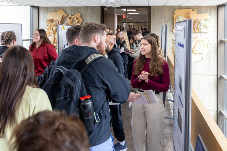 A student talks to a crowd about their research poster during poster presentations on the 4th-floor link.