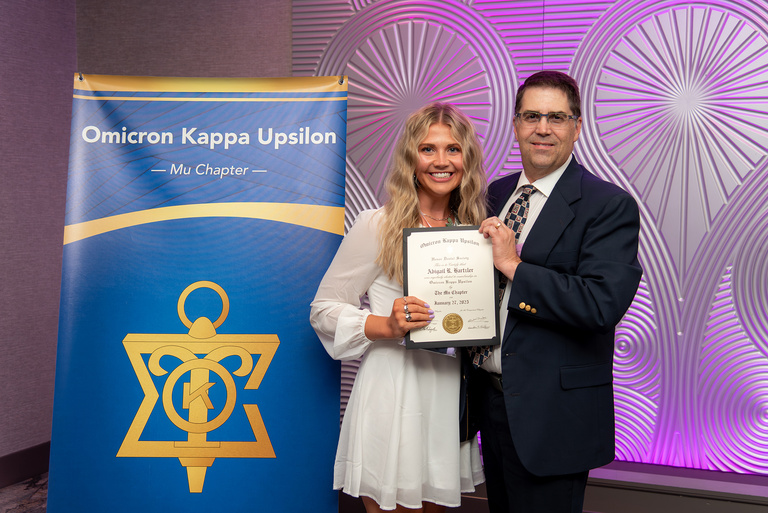 Abigail Hartzler poses for a picture in front of the OKU banner and with a certificate after being inducted into OKU.