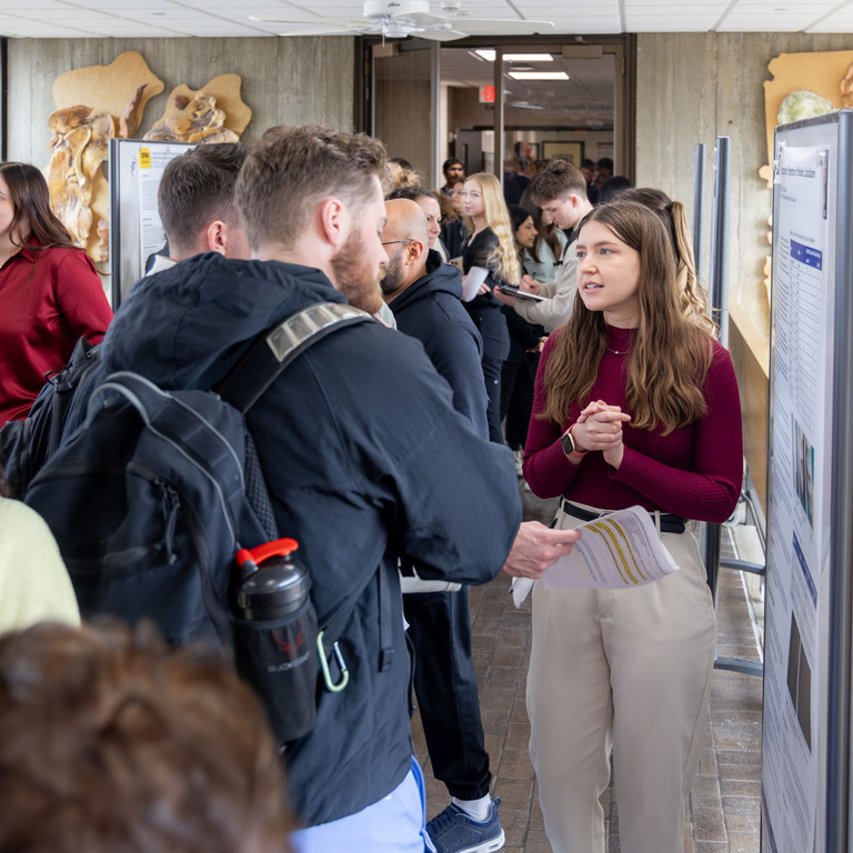 A student talks to a crowd about their research poster during poster presentations on the 4th-floor link.
